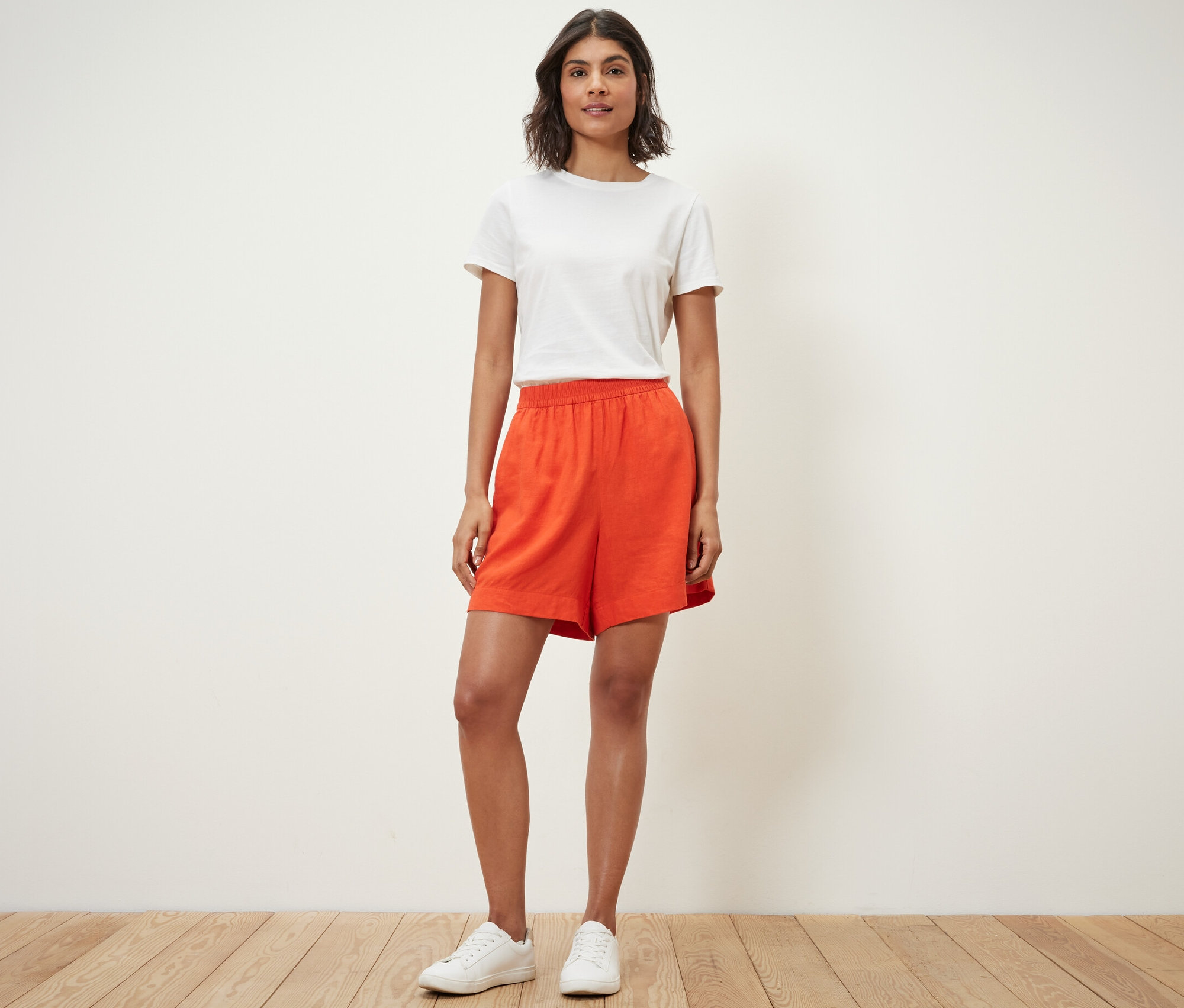Frau steht und trägt Leinenshorts, orange, und ein weißes T-Shirt.
