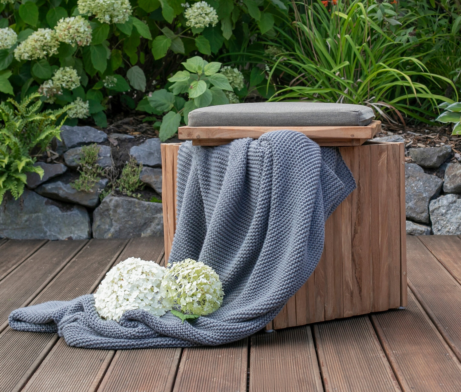 Ein DeVries Sitzhocker mit Stauraum, eine graue Decke und weiße Hortensien auf einer Holzterrasse.