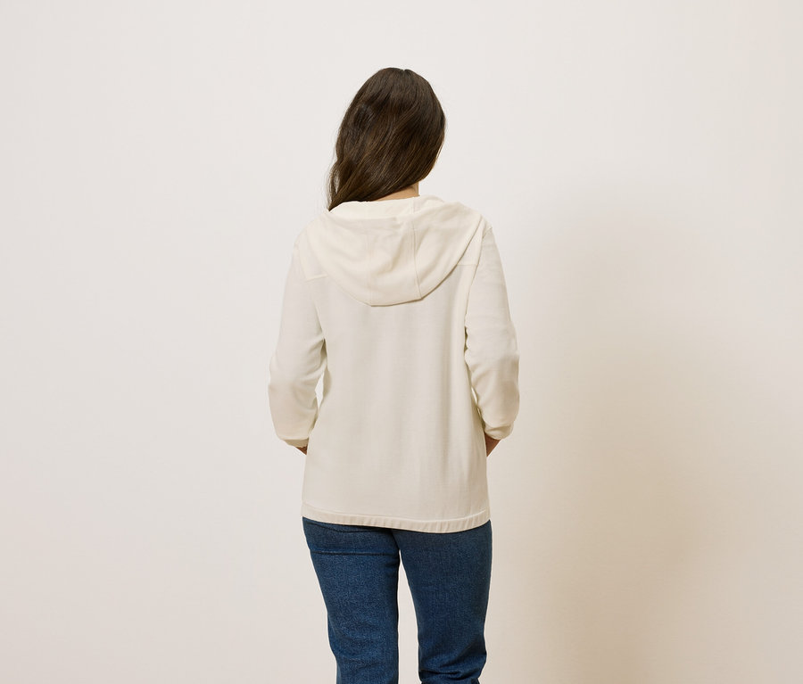 Rückansicht einer Frau mit braunen Haaren, die einen cremefarbenen Kapuzenpullover und blaue Jeans trägt.