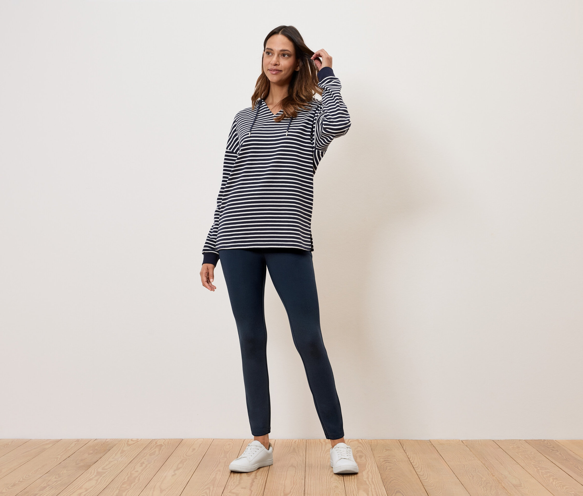 Eine Frau steht auf einem Holzboden und trägt ein gestreiftes Oberteil, dunkelblaue Leggings und weiße Turnschuhe.