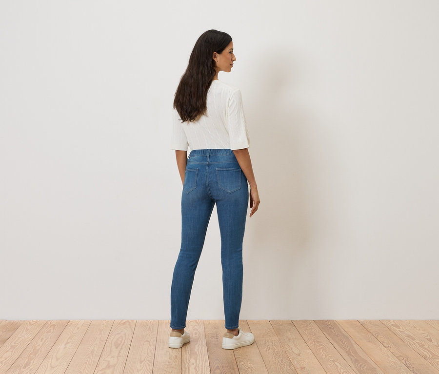 Eine Frau steht mit dem Rücken zur Kamera und trägt blaue Jeans, ein weißes Oberteil und weiße Turnschuhe.