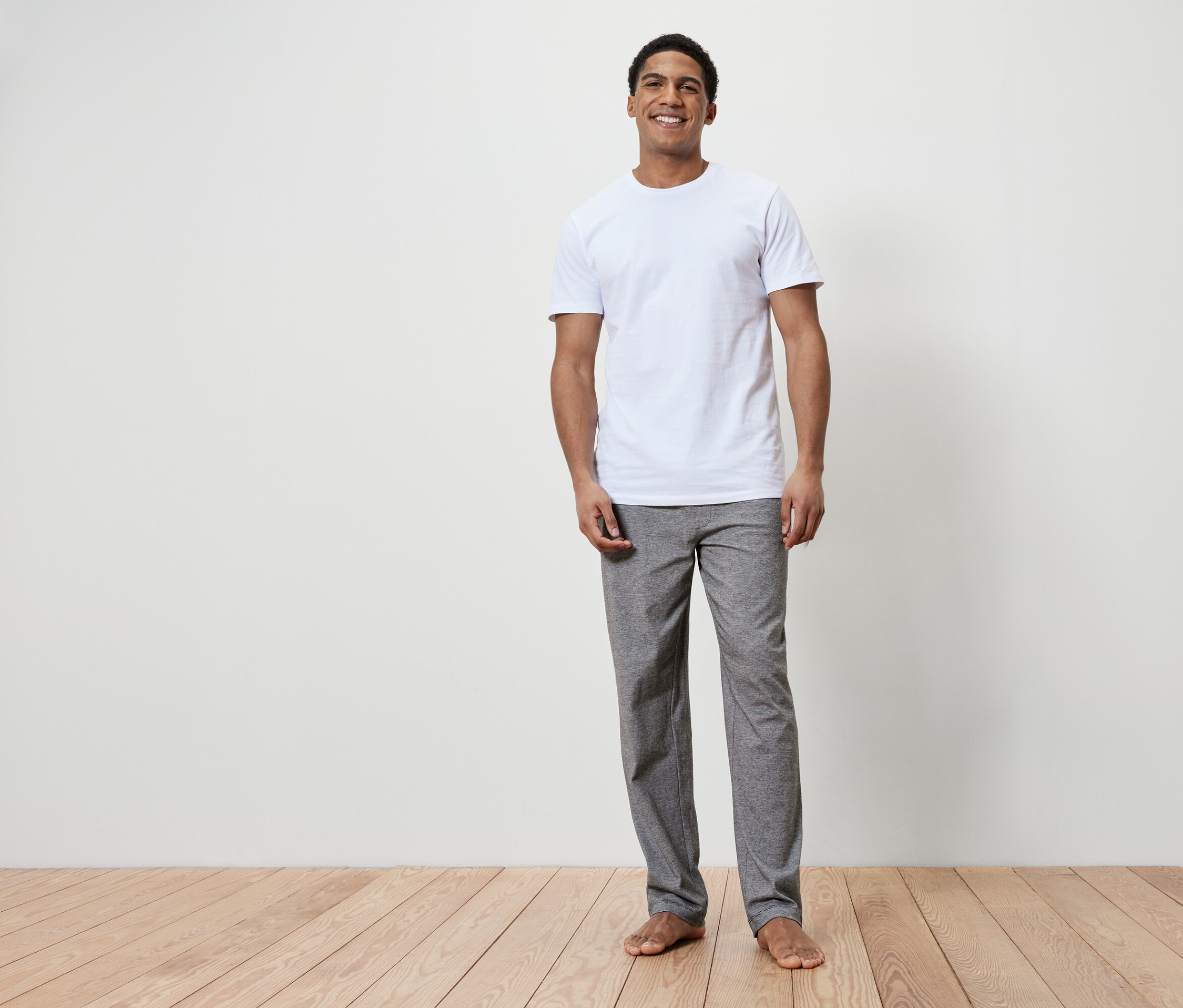 Ein lächelnder Mann steht barfuß auf einem Holzboden. Er trägt ein weißes T-Shirt und graue Hosen.