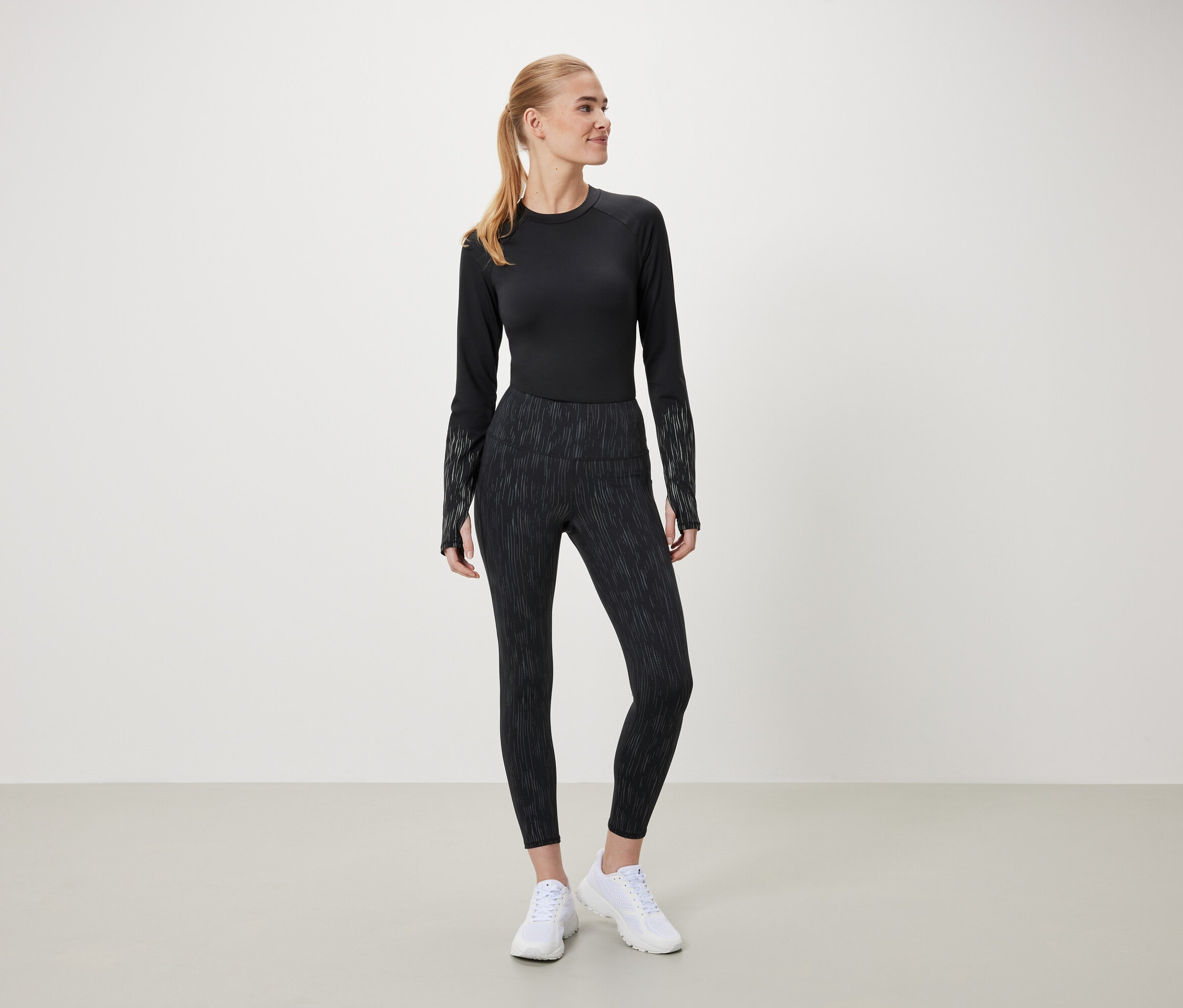 Eine Frau in einem schwarzen Langarmshirt und gemusterten Leggings steht vor einem hellen Hintergrund.