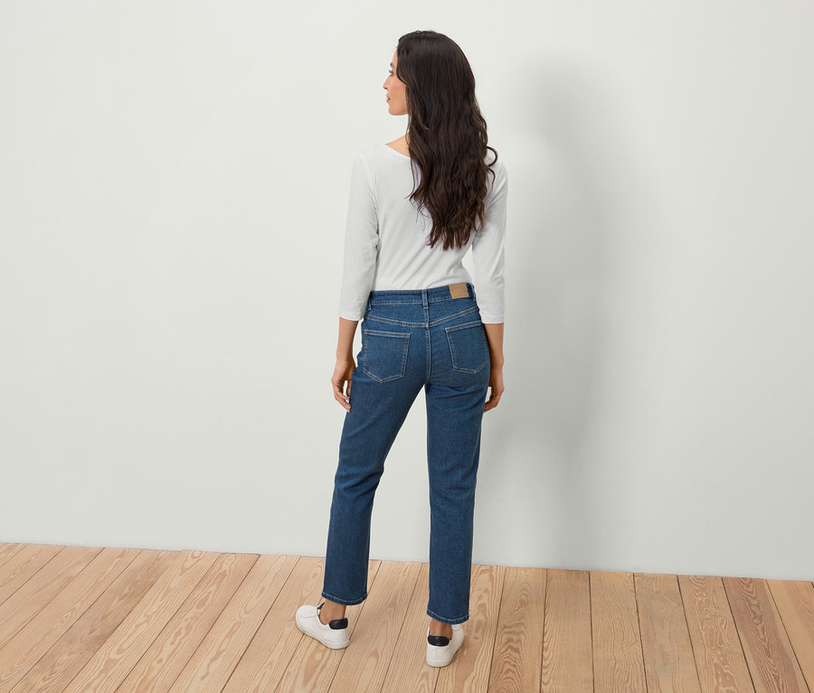 Frau mit langen braunen Haaren steht mit dem Rücken zur Kamera in blauen Jeans und einem weißen Oberteil.