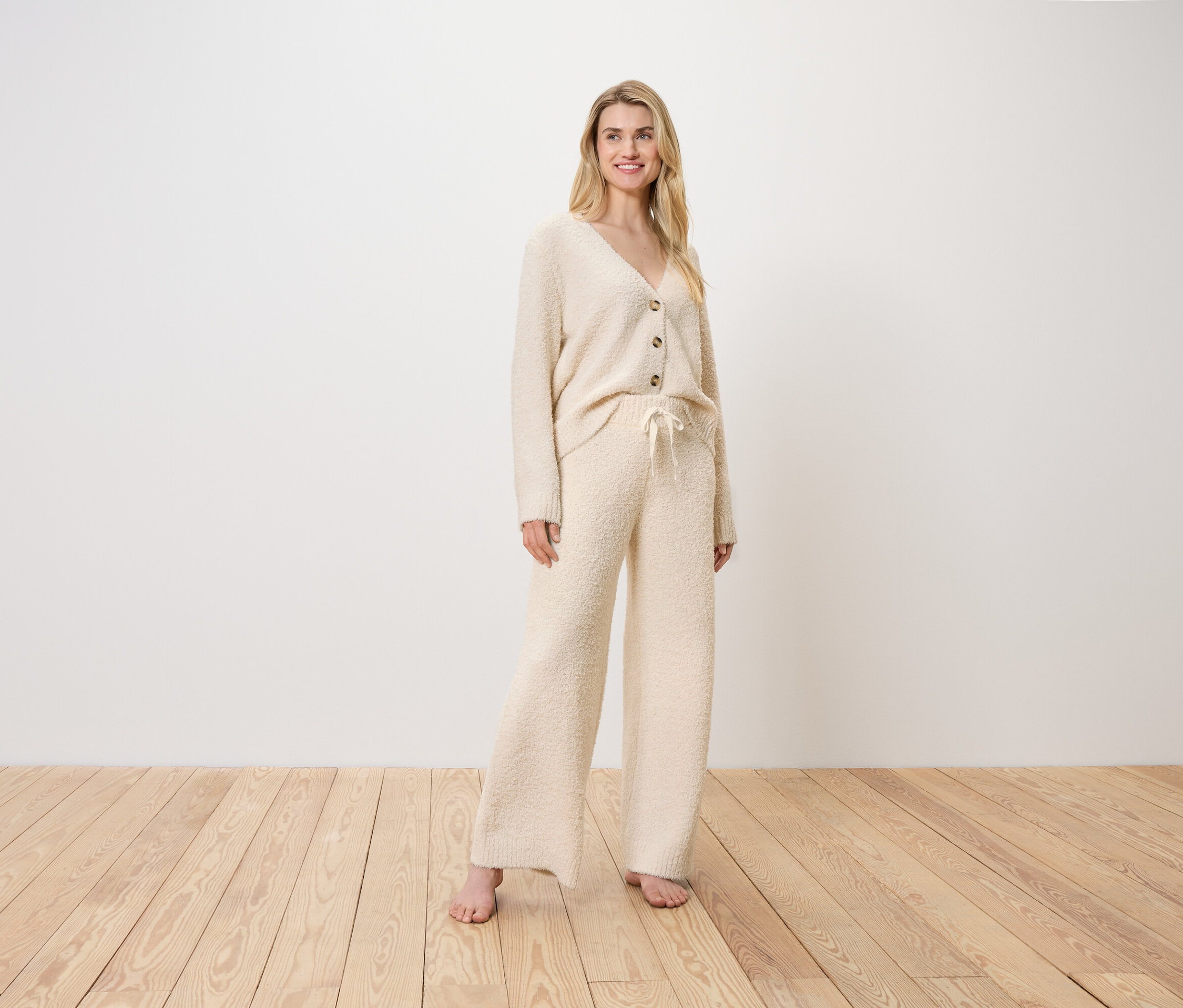 Blonde Frau steht in einem cremefarbenen Cardigan und Hose auf einem Holzboden.