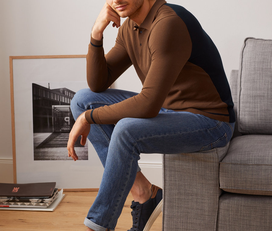 Mann sitzt auf Couch und trägt ein Strick-Poloshirt, Jeans und Turnschuhe.