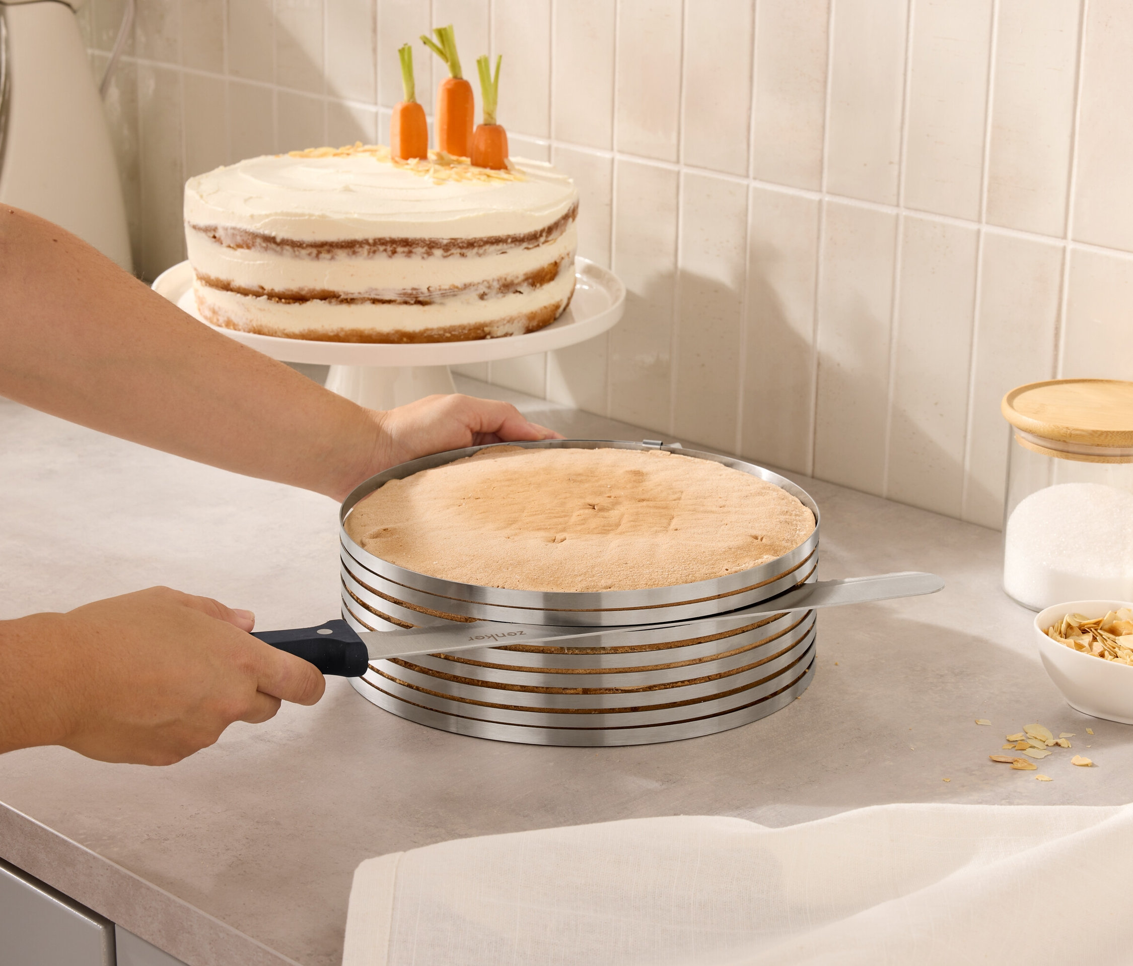 Eine Person schneidet einen Kuchen mit einem verstellbaren Kuchenschneider. Im Hintergrund befinden sich ein Kuchen mit Karotten und Gläser mit Deckeln.