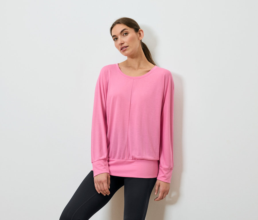 Frau posiert in einem pinkfarbenen, langärmeligen Sportoberteil und schwarzen Leggings.