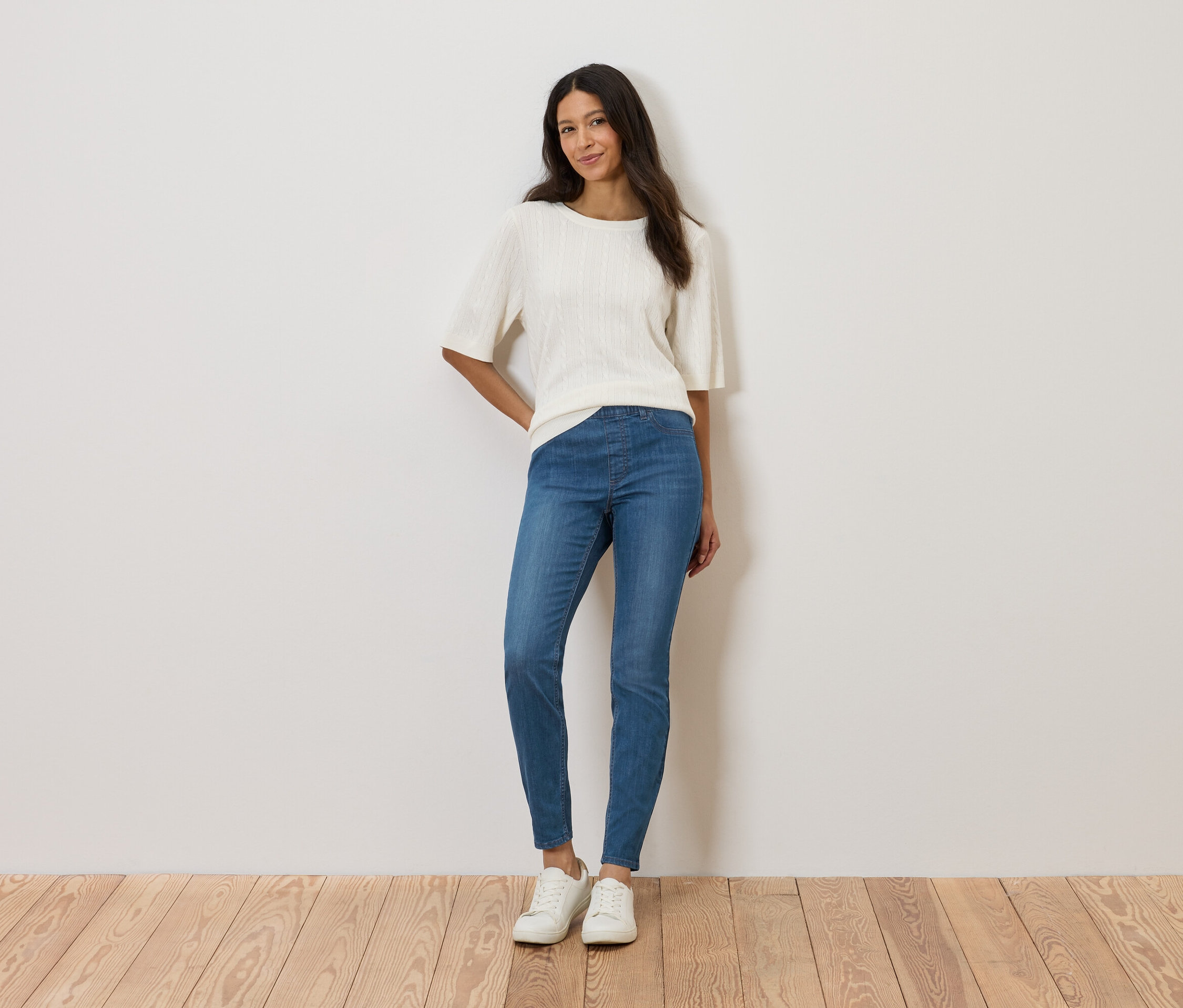 Frau steht vor einer Wand und trägt ein weißes Oberteil, blaue Jeans und weiße Schuhe.
