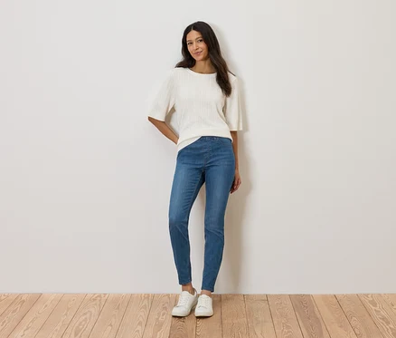 Frau steht vor einer Wand und trägt ein weißes Oberteil, blaue Jeans und weiße Schuhe.