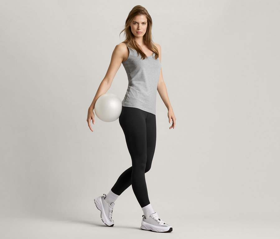 Frau hält einen Ball, trägt schwarze Leggings, Sportschuhe und 2 Tops, schwarz/grau meliert.