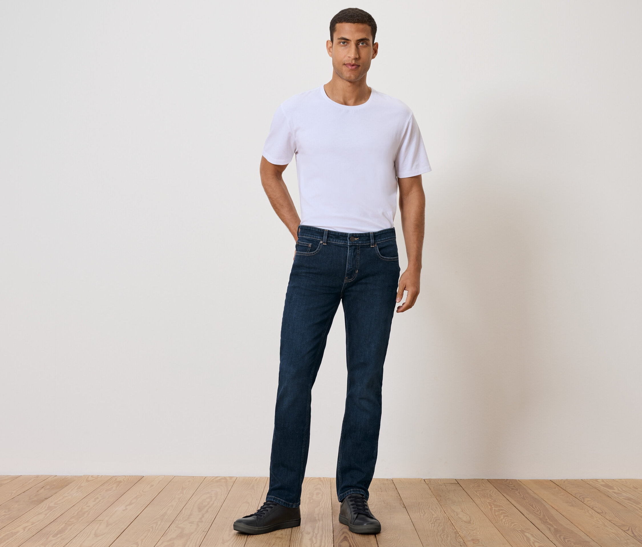 Ein Mann steht mit der Hand in der Tasche und trägt ein weißes T-Shirt, Jeans und schwarze Schuhe.