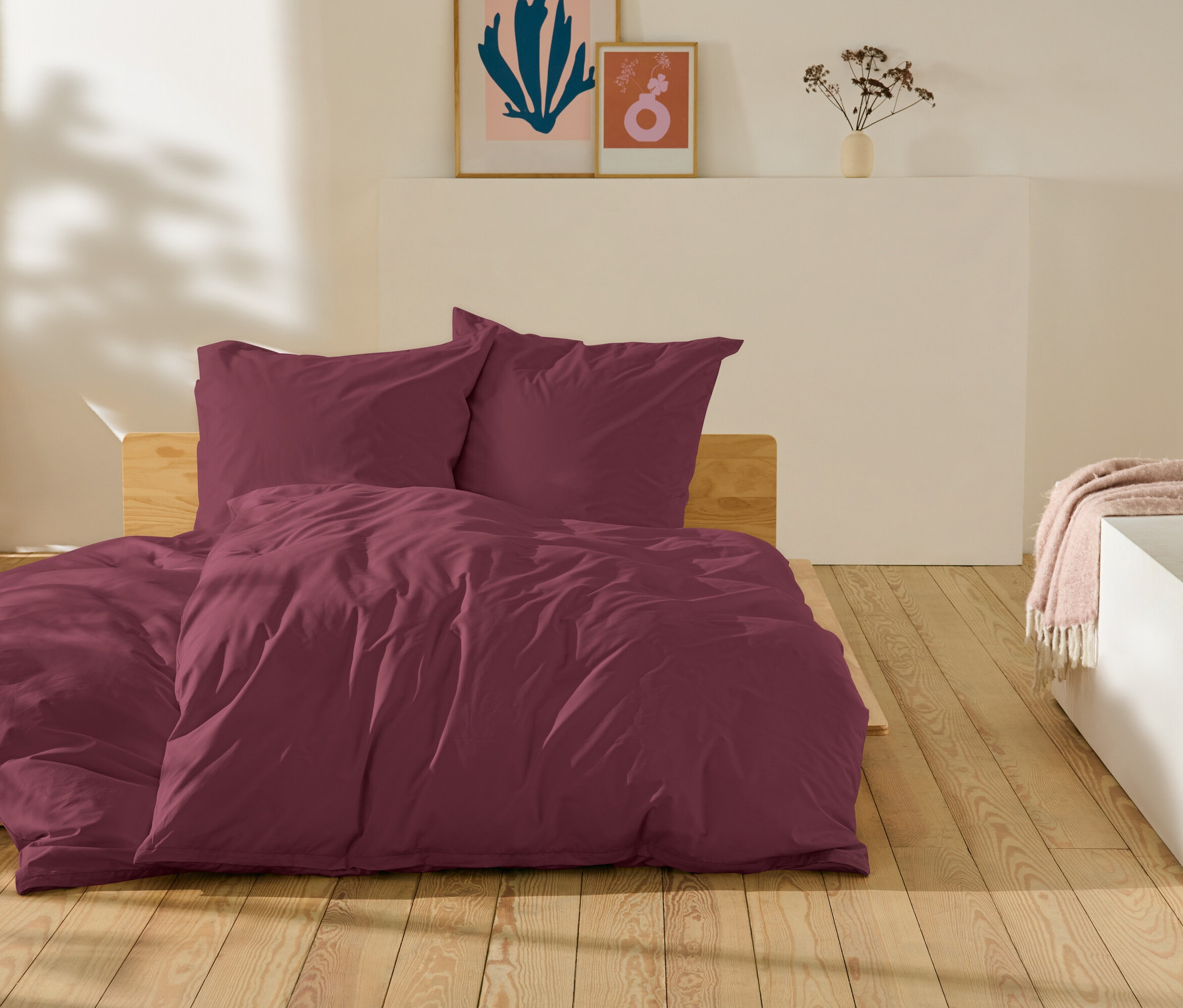 Weinrote Bettwäsche liegt auf einem Bett auf einem Holzboden.