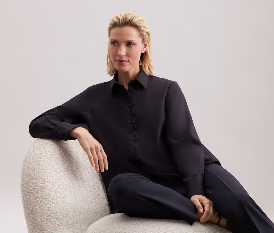 Eine Frau sitzt in einem Sessel und trägt ein schwarzes Hemd und eine Hose.