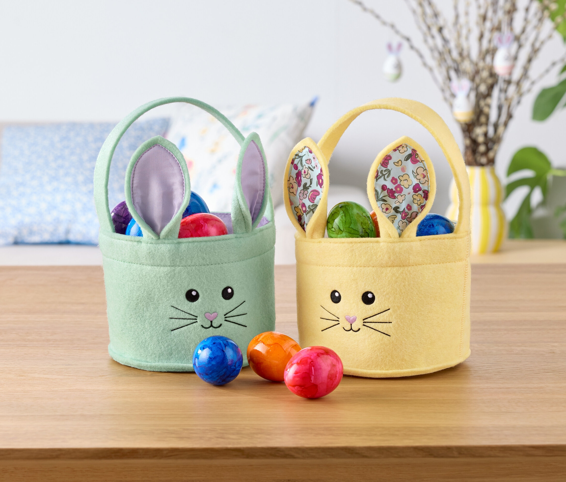 Zwei österliche Hasenkörbchen, eines mintgrün und eines gelb, gefüllt mit bunten Eiern, stehen auf einem Tisch.