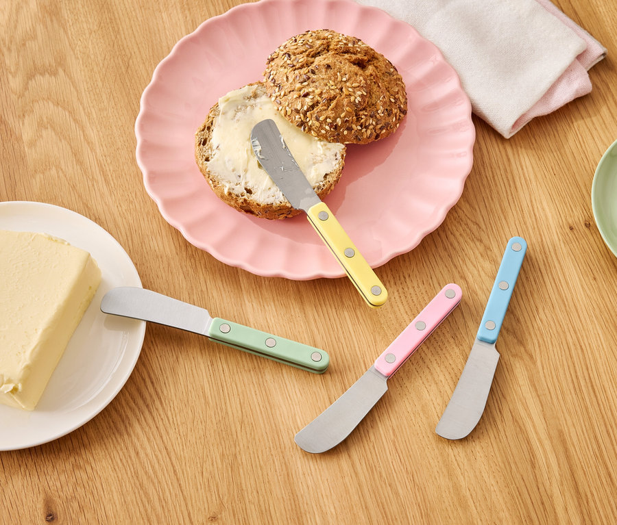Ein Brötchen mit Butter auf einem rosa Teller neben einem Stück Butter und bunten Messern auf einem Holztisch.