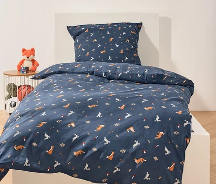 Auf einem weißen Bett liegt marineblaue Bettwäsche mit einem Muster aus Füchsen, Hasen, Igeln und Pilzen. Neben dem Bett steht ein Tisch mit einem Plüschfuchs und Spielzeug.