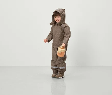 Ein lächelndes Kind in einer braunen, bedruckten Jacke mit Kapuze und Hose hält eine orangefarbene Gießkanne.
