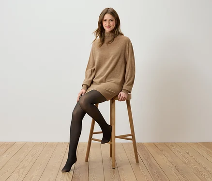 Frau sitzt auf einem Holzhocker, trägt ein braunes Rollkragenkleid und eine schwarze Strumpfhose.
