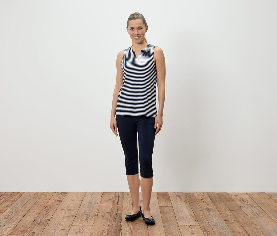 Eine Frau steht vor einer weißen Wand und trägt ein blau-weiß gestreiftes Tanktop, blaue Caprihosen und Ballerinaschuhe.