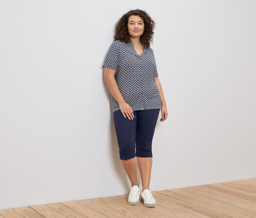 Eine Frau mit lockigem Haar lehnt an einer Wand und trägt eine blau-weiß gemusterte Bluse, blaue Caprihosen und weiße Turnschuhe.