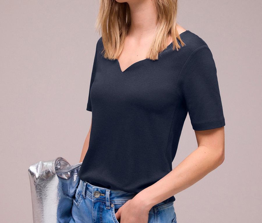 Junge blonde Frau lächelt, trägt ein marineblaues Street One Kurzarmshirt und hellblaue Jeans, hält eine silberne Tasche.