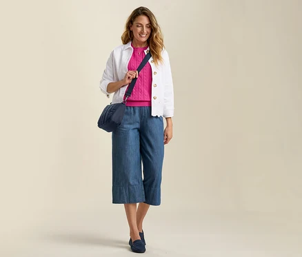 Frau mit Jeans-Culottes, pinkem Pullover und Jeansjacke mit Lochstickerei. Blaue Umhängetasche und passende Slipper.