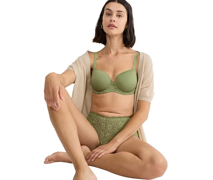 Frau sitzt mit gekreuzten Beinen und trägt einen grünen BH und eine grüne Spitzhose, zusammen mit einer beigen Strickjacke.