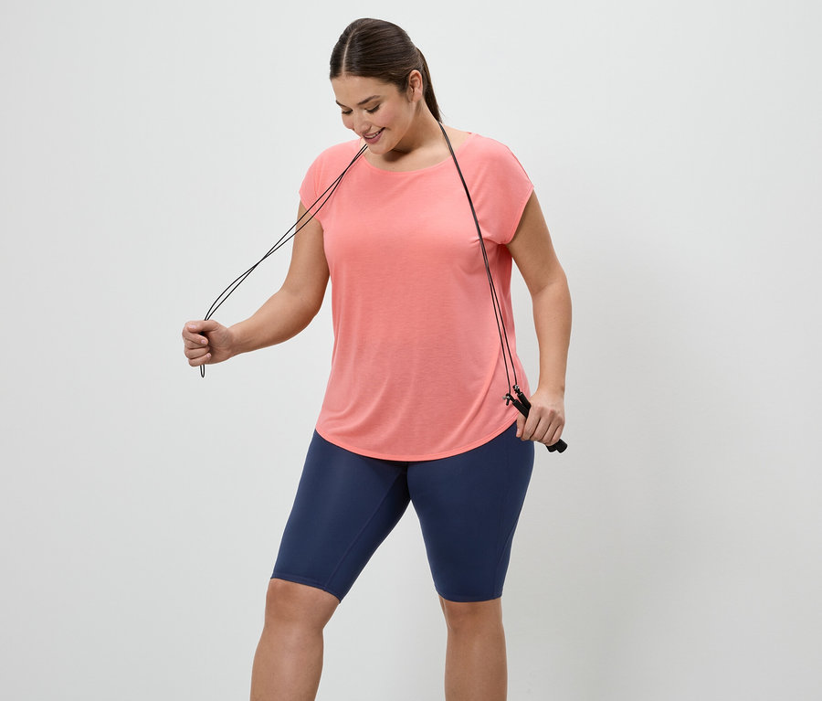 Frau in lachsfarbenem T-Shirt und blauen Radlerhosen hält ein Springseil vor einem weißen Hintergrund.