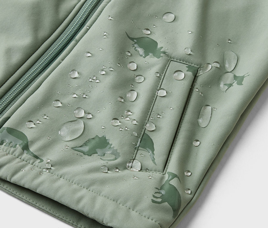 Nahaufnahme einer hellgrünen Jacke mit Reißverschluss, Tasche und Dinosaurier-Muster, bedeckt mit Wassertropfen.