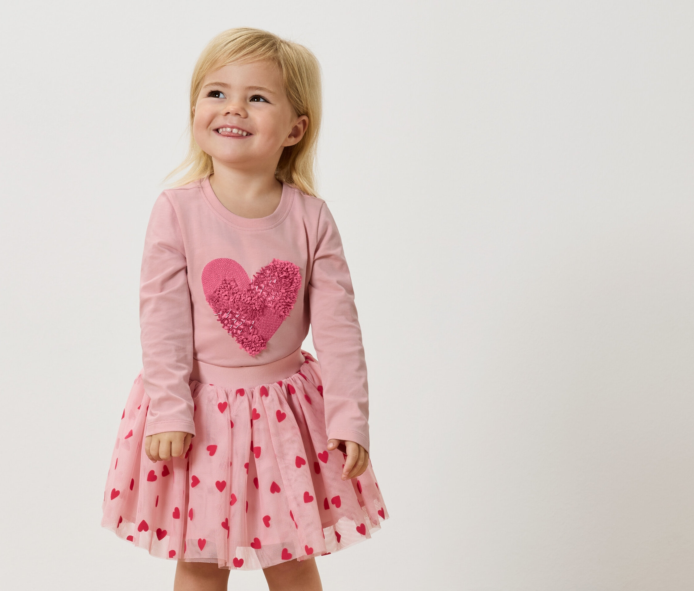 Ein kleines Mädchen mit blonden Haaren trägt ein rosa Langarmshirt mit einem großen, rosafarbenen Herz und einen rosa Tüllrock mit roten Herzen.