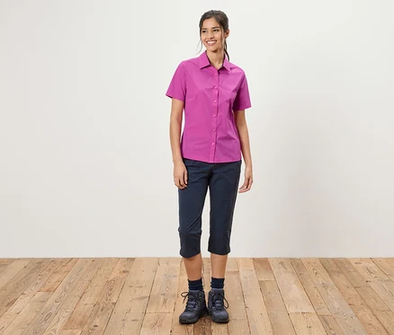 Eine Frau steht vor einer weißen Wand. Sie trägt ein pinkes Hemd, blaue Dreiviertelhosen und Wanderschuhe.