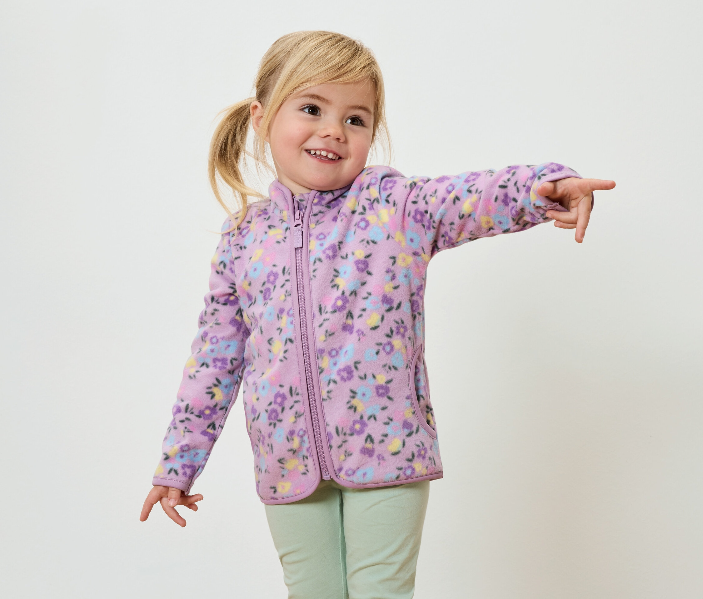 Ein lächelndes Mädchen mit blonden Zöpfen, bekleidet mit einer lila Fleecejacke mit Blumenmuster und hellgrünen Hosen, zeigt mit dem Finger.
