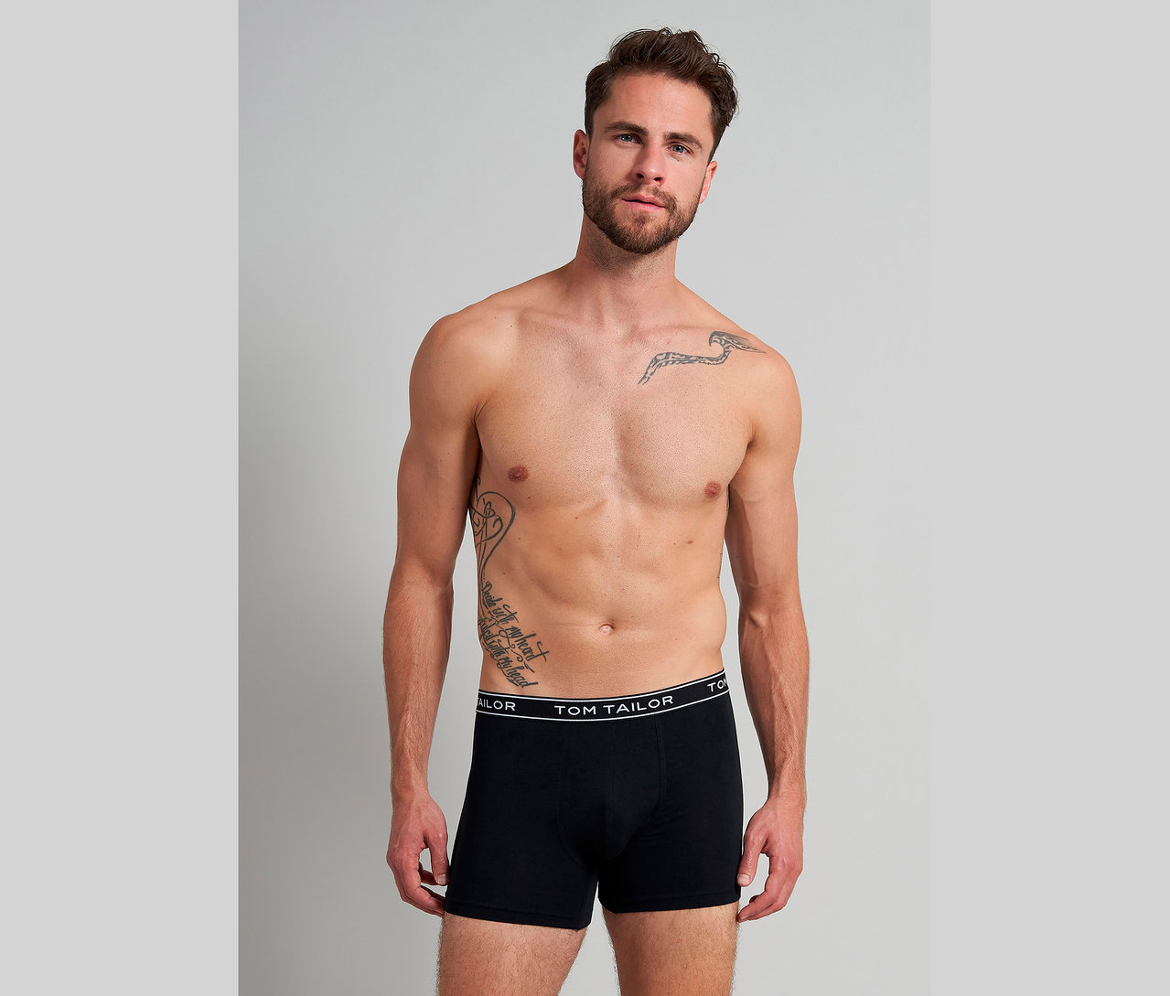 Mann mit freiem Oberkörper und Tätowierungen, der schwarze TOM TAILOR Boxershorts trägt.