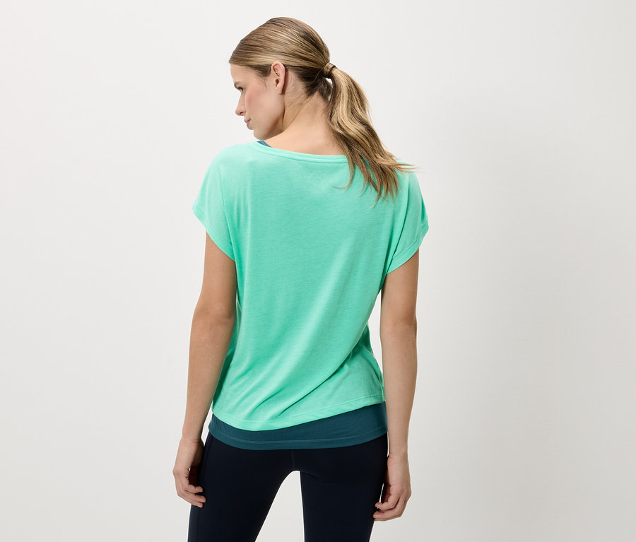 Rückansicht einer Frau mit blonden Haaren, die ein grünes T-Shirt und dunkle Leggings trägt.