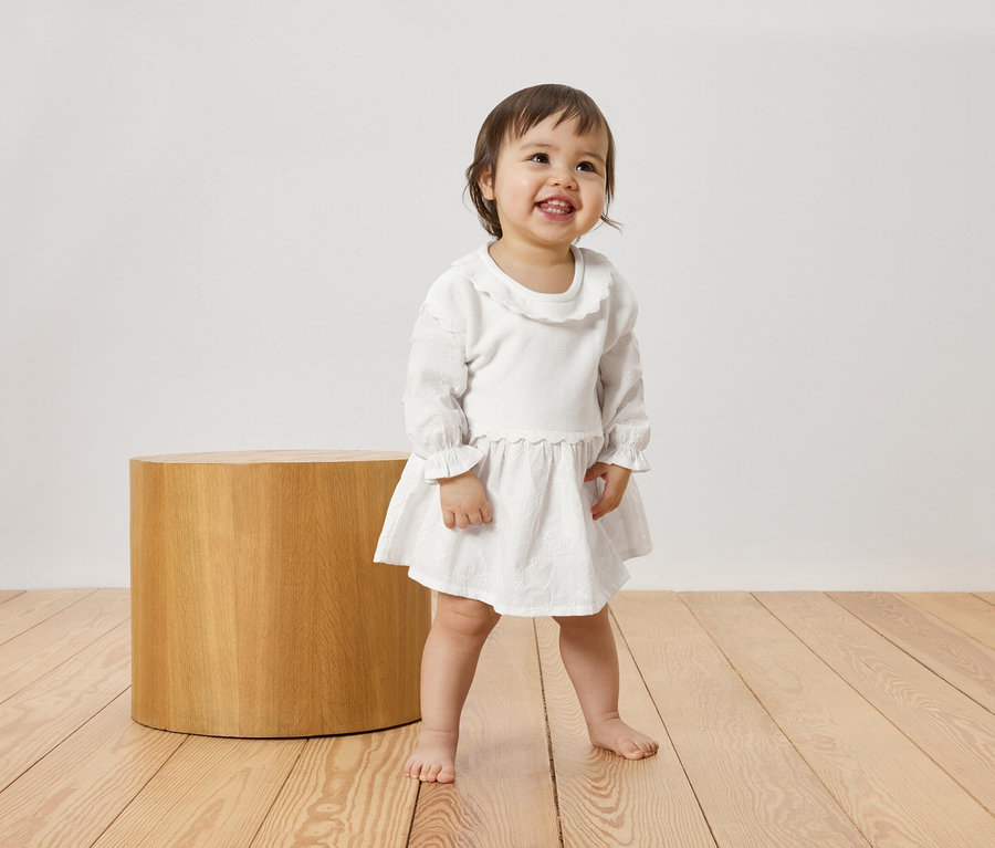 Ein barfüßiges, lachendes Kleinkind steht auf einem Holzboden neben einem zylindrischen Hocker und trägt ein weißes Kleid.