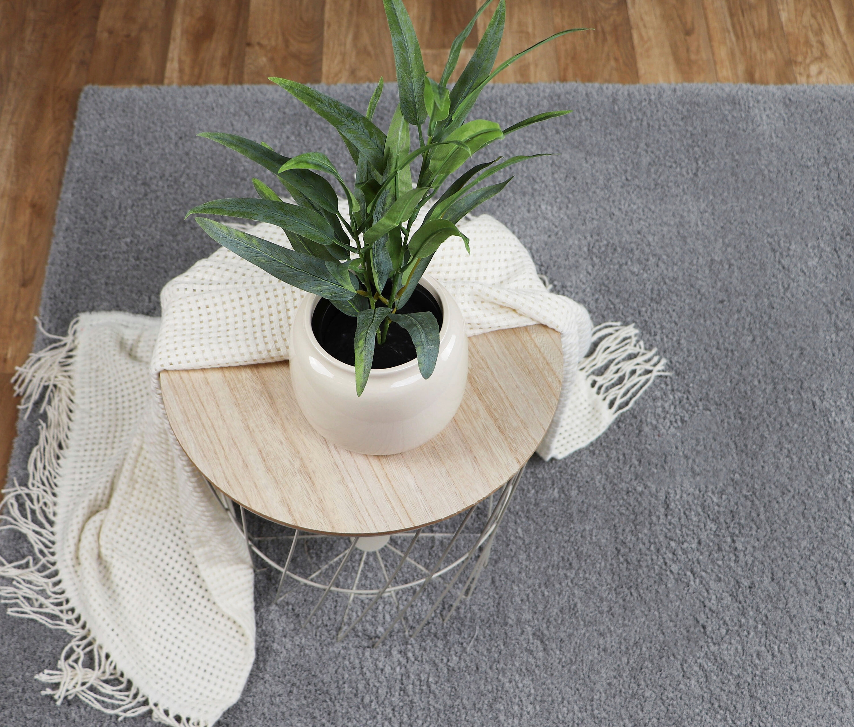 Auf einem hellgrauen »Pleasure« Teppich steht ein Tisch mit einer Pflanze und einer weißen Decke.