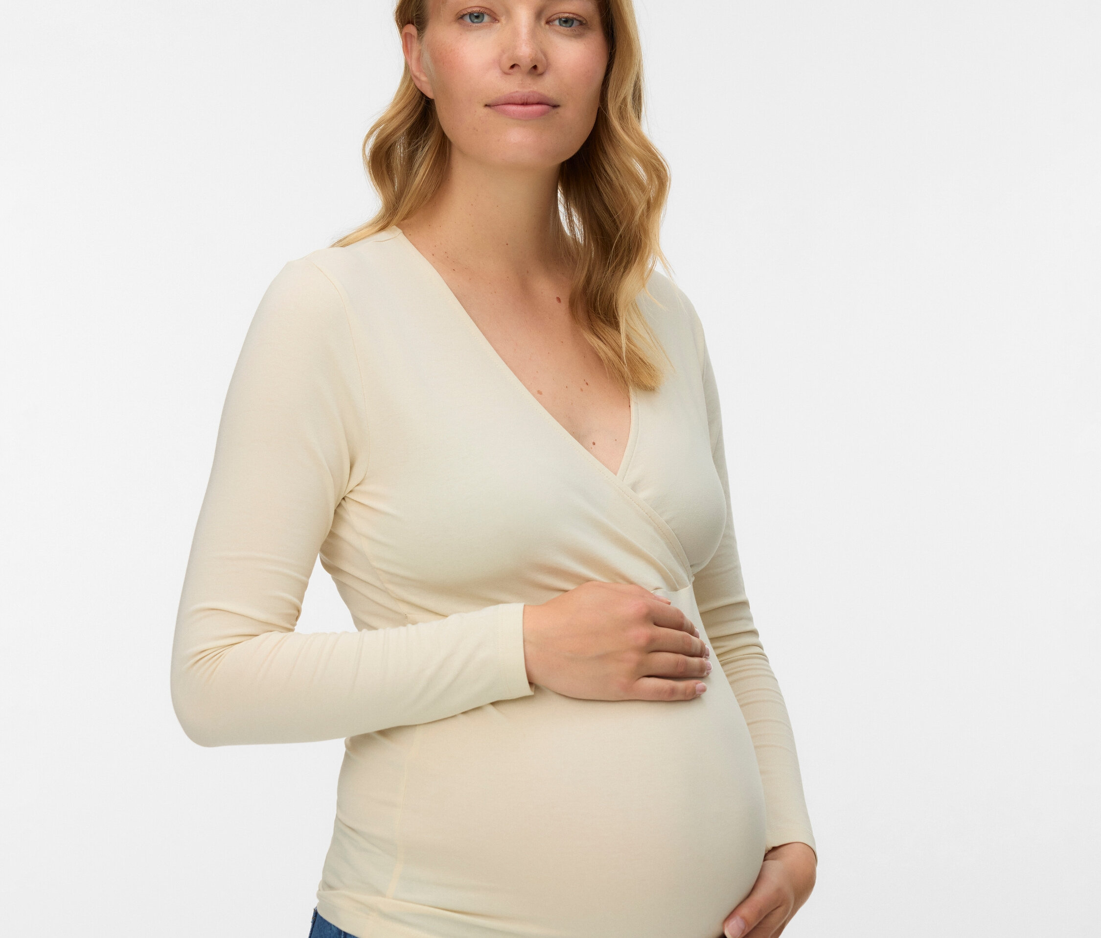 Eine schwangere Frau mit blonden, gewellten Haaren trägt eine Wickelbluse mit langen Ärmeln und Jeans.