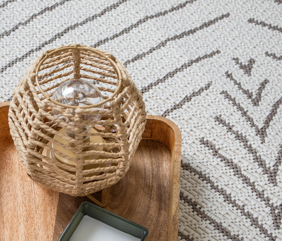 Nahaufnahme einer geflochtenen Lampe auf einem Holztablett, daneben eine Kerze, auf einem Teppich mit geometrischem Muster.