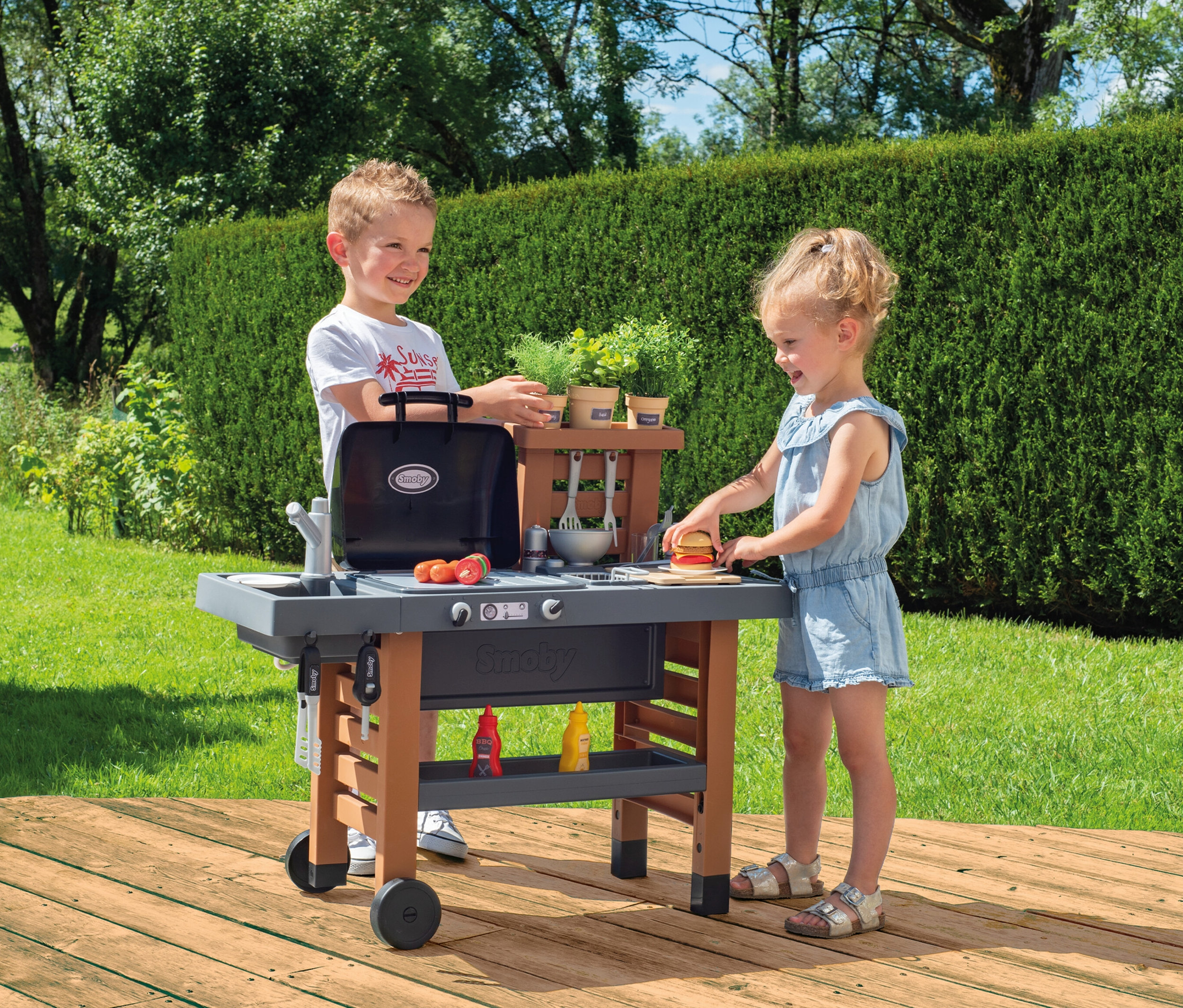 Junge und Mädchen spielen mit der Smoby Outdoor-Gartenküche im Garten.