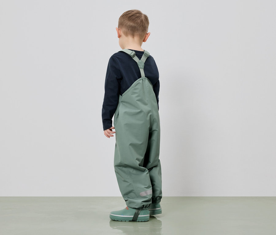 Kind von hinten in grüner Latzhose mit Gummistiefeln und blauem Shirt vor weißem Hintergrund.