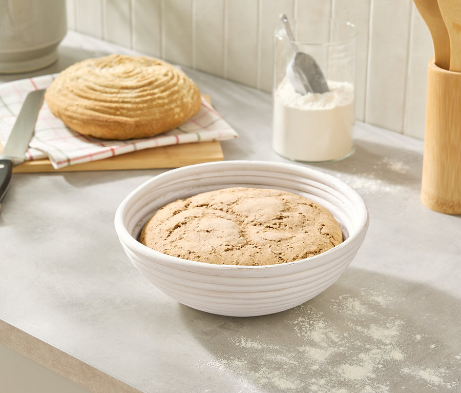 Auf einer Küchentheke stehen ein Brotlaib in einem weißen Gärkorb und ein Brot auf einem Brett neben einem Messer und einem Glas mit Mehl.
