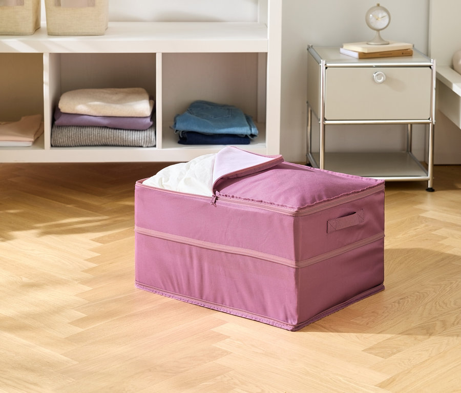 Ein rosa Stoffbehälter steht auf einem Holzboden, teilweise mit weißer Wäsche gefüllt. Im Hintergrund sind ein Regal und ein Nachttisch zu sehen.