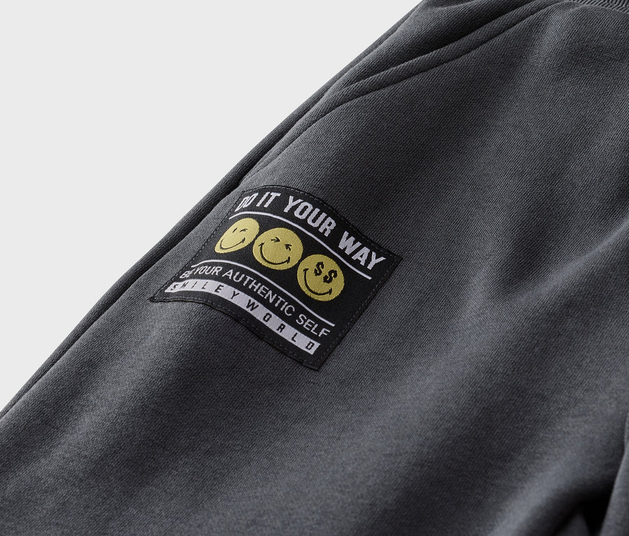 Nahaufnahme einer grauen Jogginghose mit einem schwarzen Aufnäher mit gelben Smileys und dem Text "Do it your way".