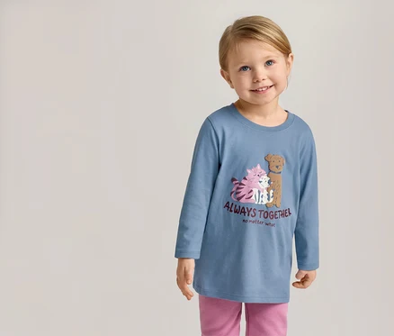 Lächelndes Mädchen mit blauem Langarmshirt mit Katzen- und Hundemotiv und dem Schriftzug "Always Together" und rosafarbener Hose.