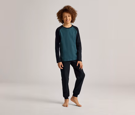 Ein lächelnder, barfüßiger Junge mit lockigem Haar posiert in einem grün-schwarzen Langarmshirt und einer schwarzen Hose vor einem hellen Hintergrund.