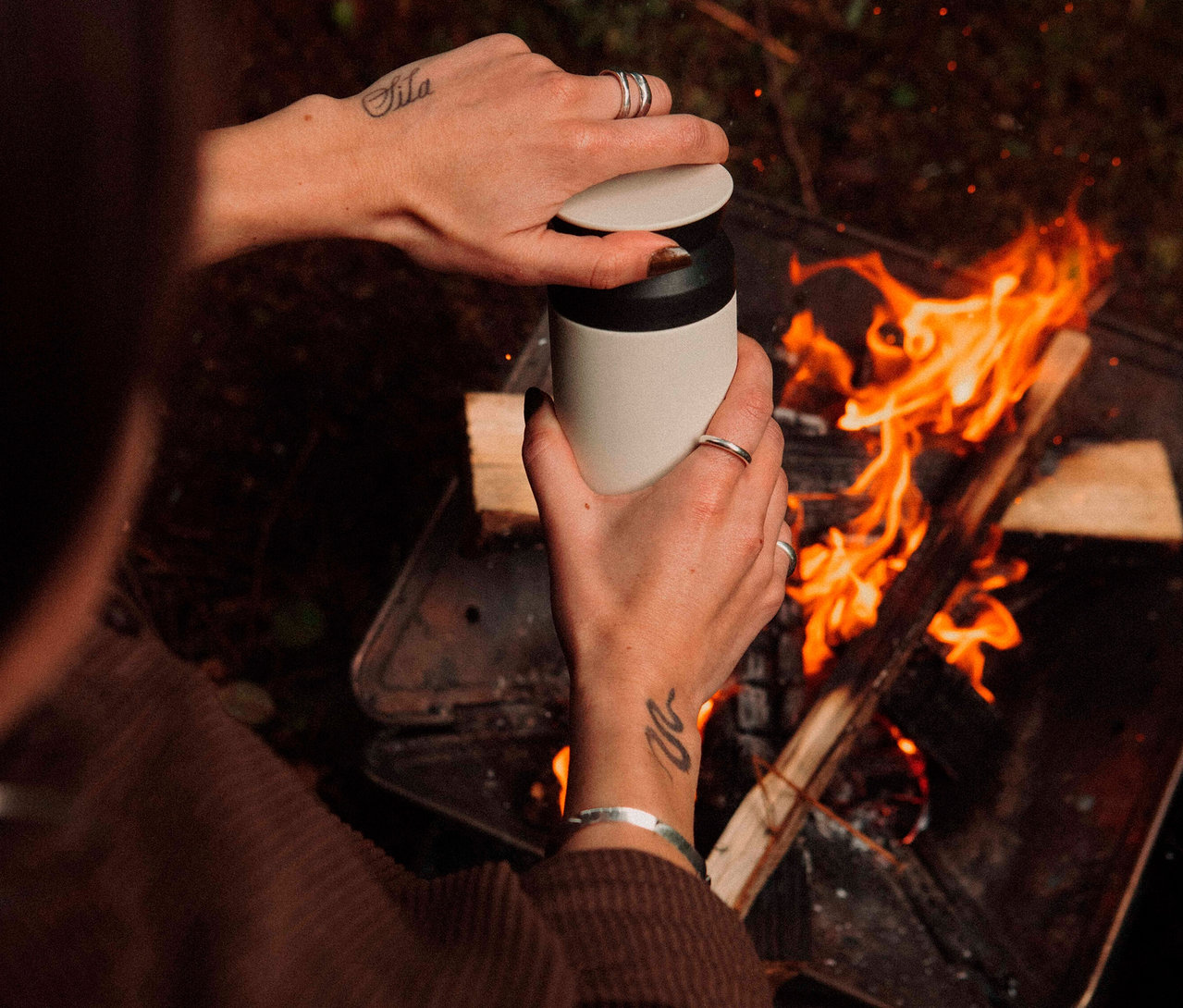 Eine Person hält eine Thermoskanne über ein Lagerfeuer.