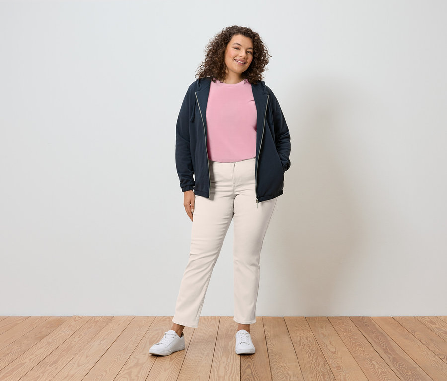 Eine Frau mit lockigem Haar steht vor einer Wand und trägt eine marineblaue Kapuzenjacke, ein rosafarbenes T-Shirt, cremefarbene Hosen und weiße Turnschuhe.