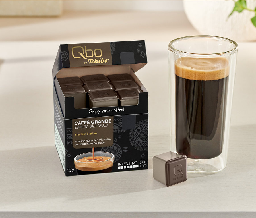 Geöffneter Karton Qbo Caffè Grande ESPÍRITO SÃO PAULO mit Kapseln und ein Glas Kaffee. Eine Kapsel liegt daneben.