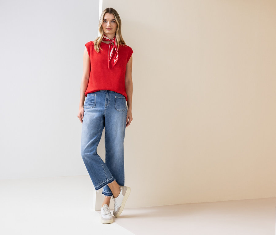 Eine Frau steht und trägt ein rotes Top, ein Halstuch, Jeans und Turnschuhe.